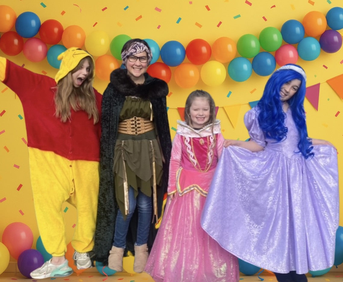 Four children in colorful costumes against a vibrant yellow wall with balloons.